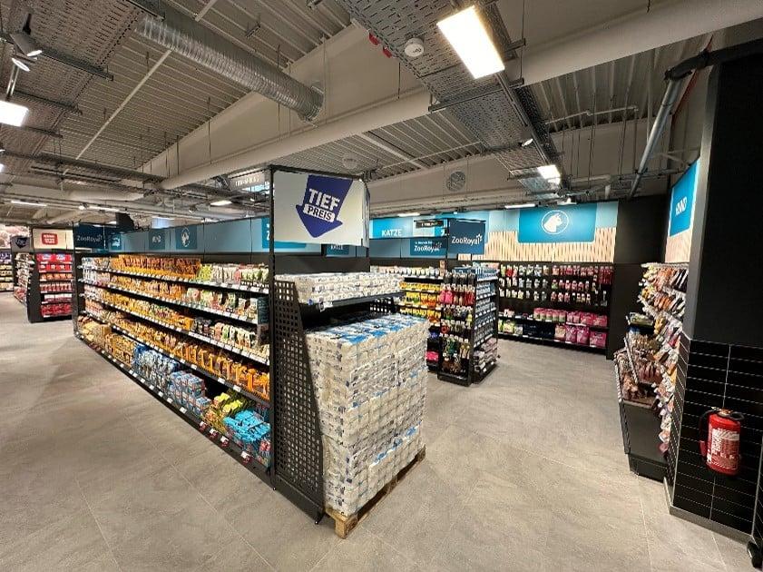 ZooRoyal Shop in Shop Heller Supermarkt‑Gang mit gut gefüllten Regalen für Snacks und Getränke, zentral gestapeltem Wasser und einem blauen „Tiefpreis“-Hinweisschild.