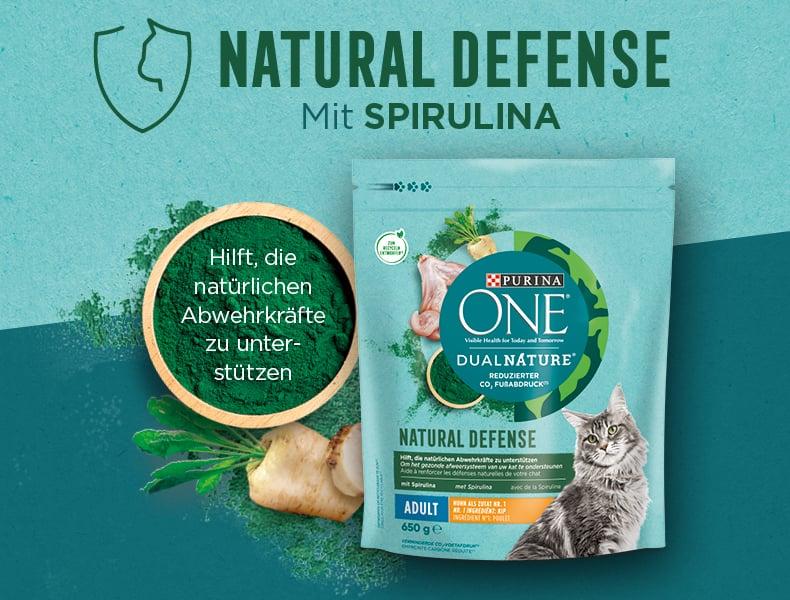 Purina ONE Natural Defense Purina‑ONE‑Natural‑Defense Produktbeutel neben Spirulina‑Zutaten und Hinweis auf Abwehrkräfte.